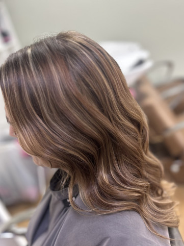 A close-up side view of a person with long, wavy hair featuring a blend of blonde and light brown highlights. The hair is styled in loose waves, and the background shows a blurred indoor setting