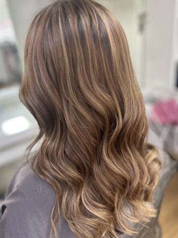 A close-up of a woman with long, wavy hair featuring a blend of blonde and light brown highlights, styled in loose waves. The photo is taken from behind, showcasing the hair's volume and texture.