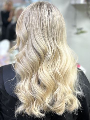 A woman with long, wavy, platinum blonde hair is shown from the back, displaying the smooth, glossy curls cascading down. The background is blurred, focusing on the hair.