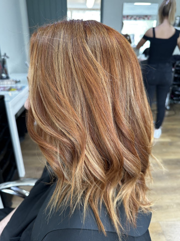 A person with shoulder-length, wavy hair dyed a rich copper colour with subtle highlights. They are seated in a salon, wearing a black cape, with salon equipment and another person in the background