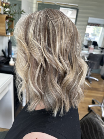 A person with short, wavy blonde hair featuring subtle highlights and lowlights, creating a natural look. They are seated in a salon, wearing a black top, with salon equipment visible in the background