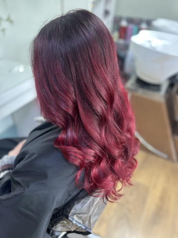 A person with long, wavy hair dyed in vibrant red, transitioning from dark roots to bright ends, seated in a salon